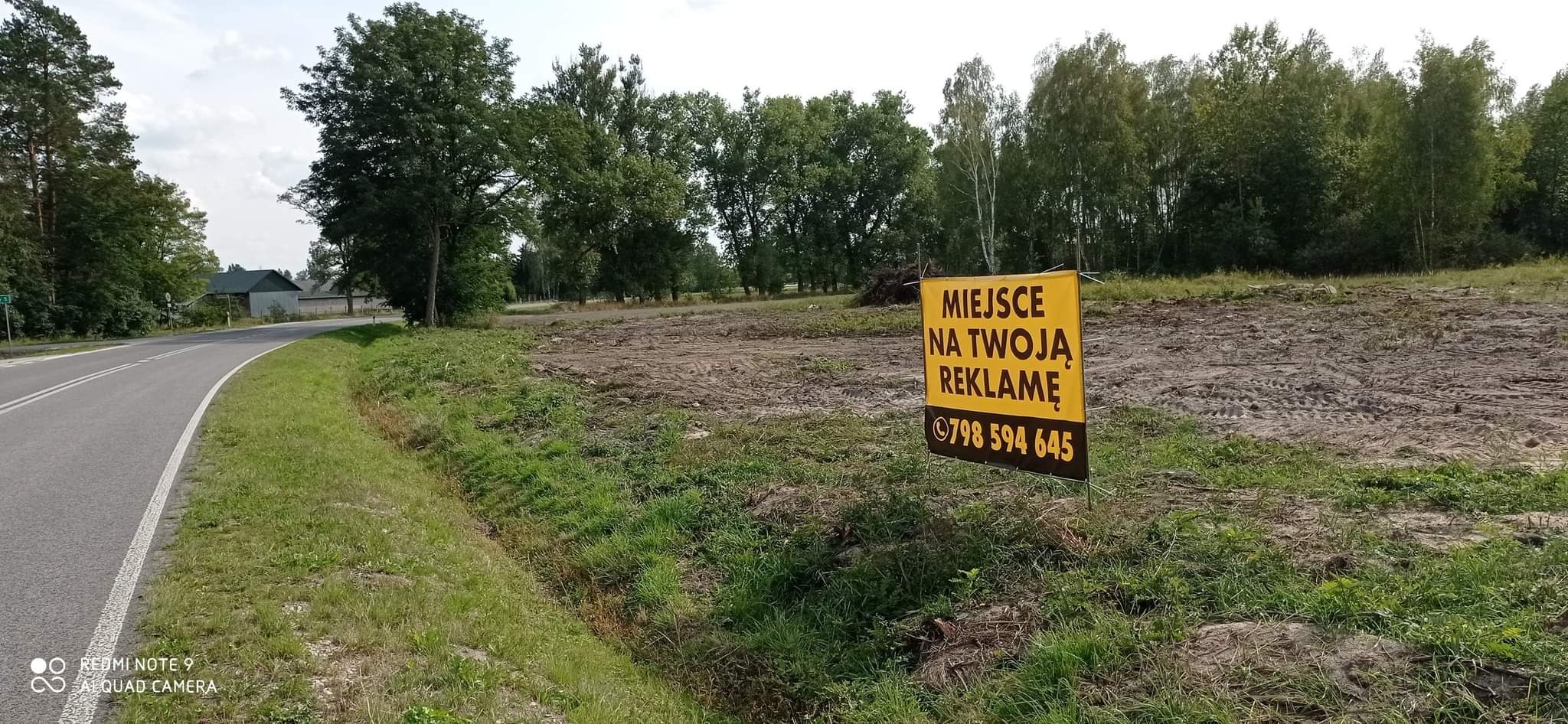Żółty baner z napisem 'Miejsce na Twoją Reklamę' i numerem telefonu na przygotowanym pod budowę terenie, widok z boku drogi.