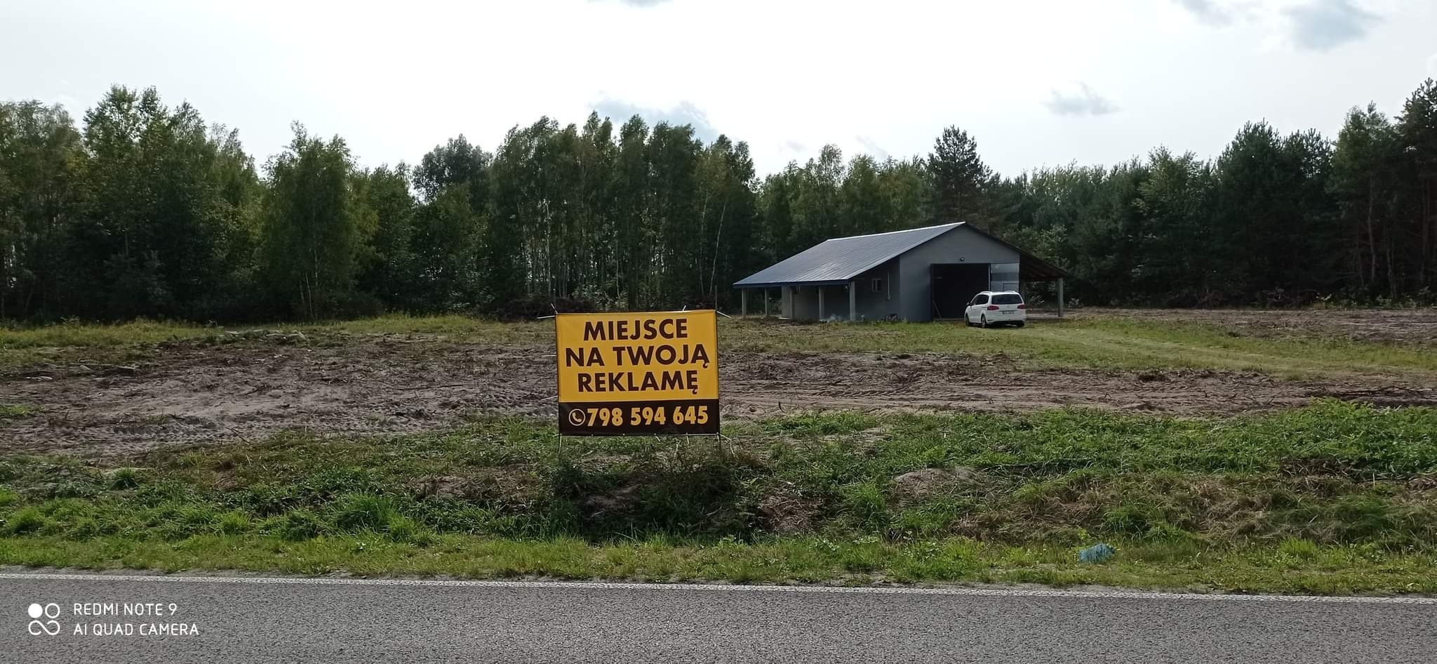 Żółty baner reklamowy z napisem 'Miejsce na Twoją Reklamę' i numerem telefonu, umieszczony na tle przygotowanego terenu z budynkiem magazynowym w oddali i lasem w tle.