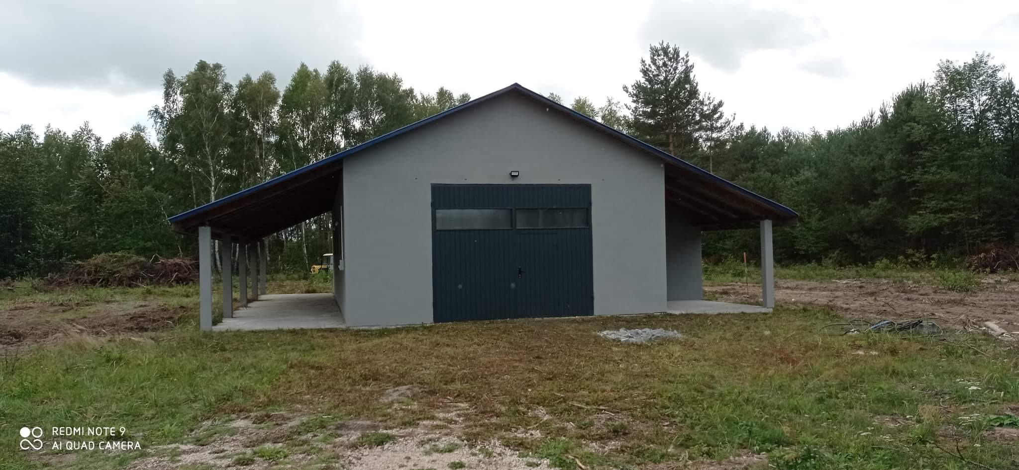 Szary budynek magazynowy z ciemnymi, metalowymi drzwiami garażowymi i zadaszonymi wjazdami po bokach, otoczony zielenią i drzewami w pochmurny dzień.