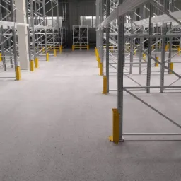 Jasnoszara, błyszcząca podłoga w magazynie z rzędami metalowych regałów zabezpieczonych żółtymi osłonami.