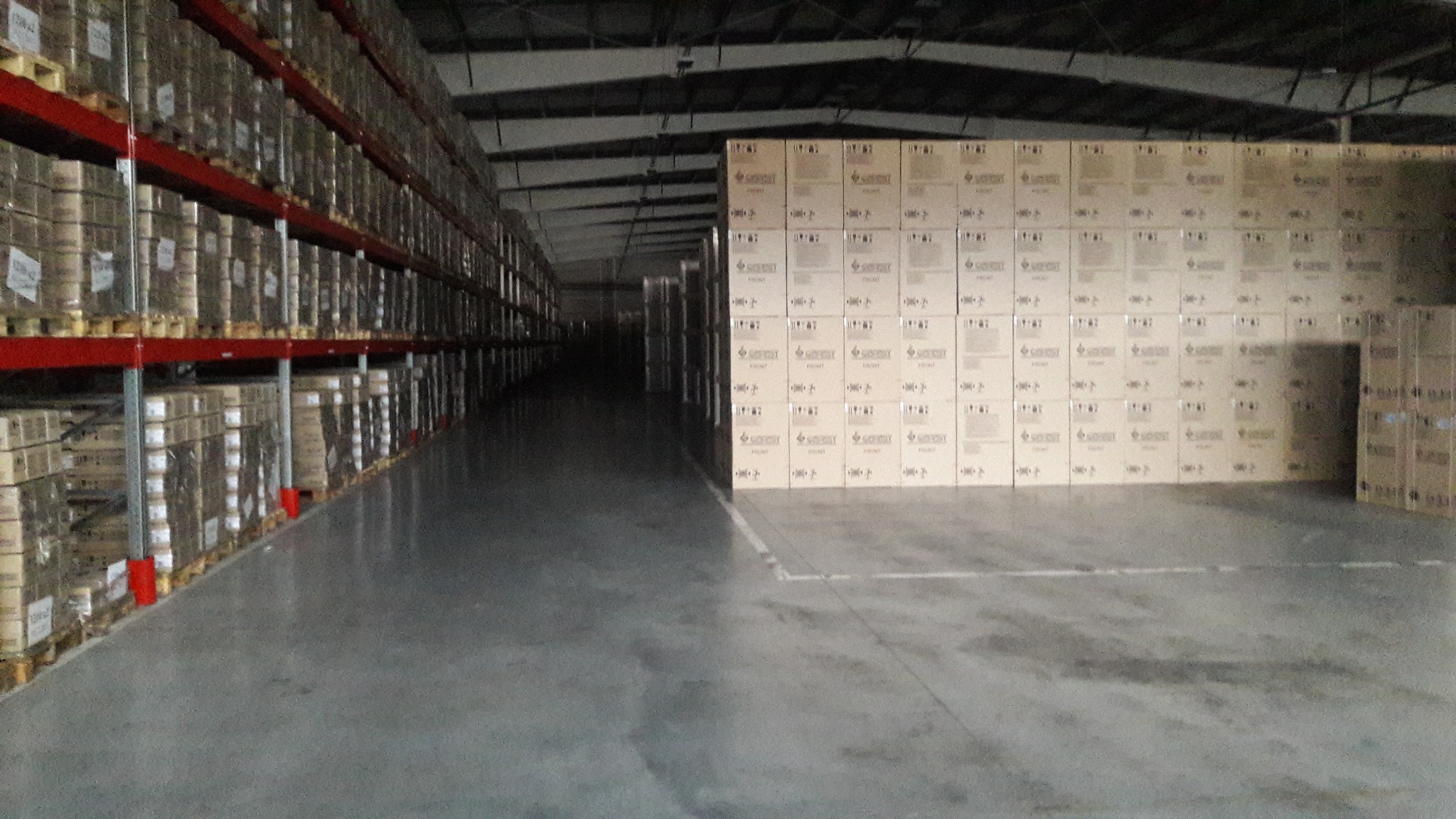 Magazyn z szarą, gładką posadzką przemysłową, regały z towarem po lewej stronie, palety z kartonami po prawej.