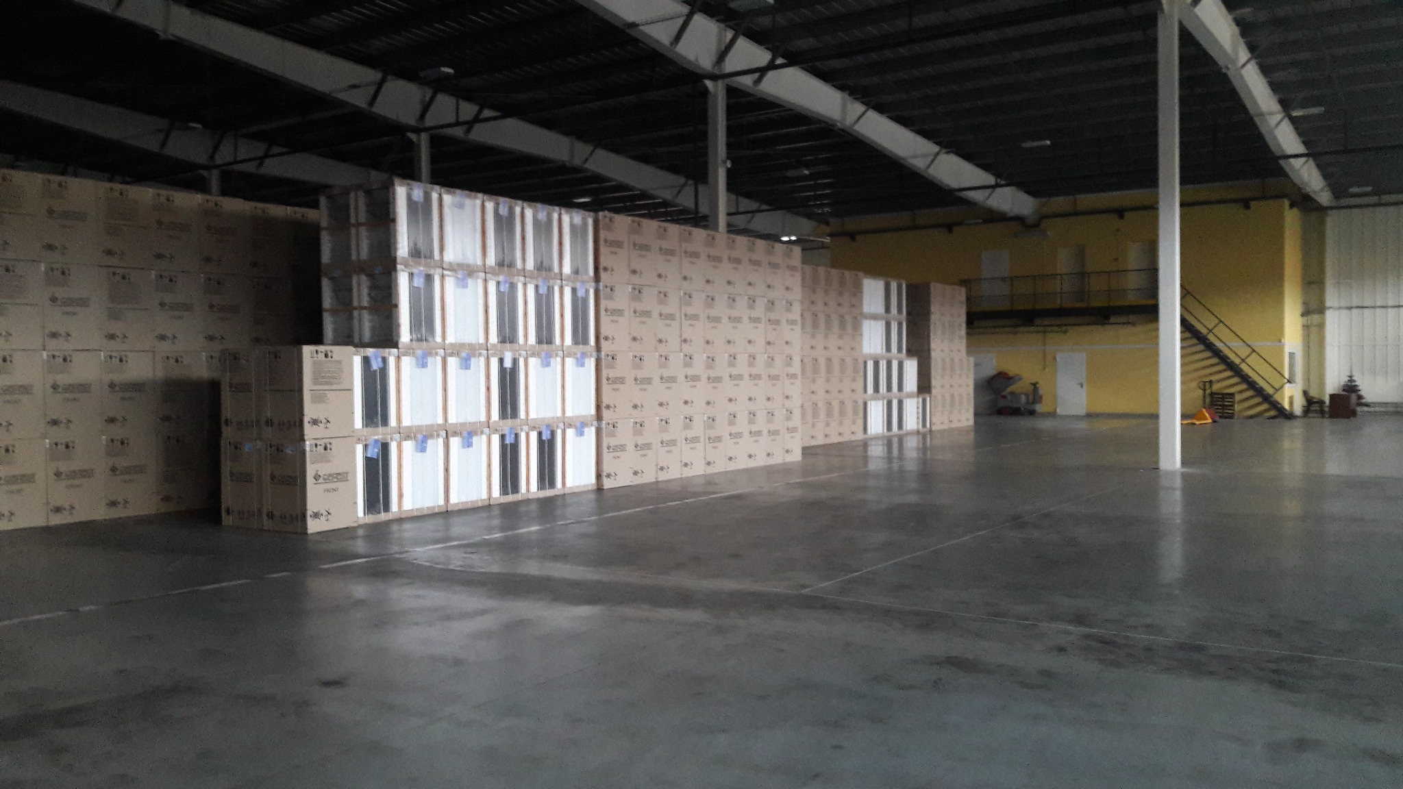 Magazyn z ułożonymi paletami kartonów na posadzce przemysłowej z widocznymi liniami podziału, żółta ściana w tle.