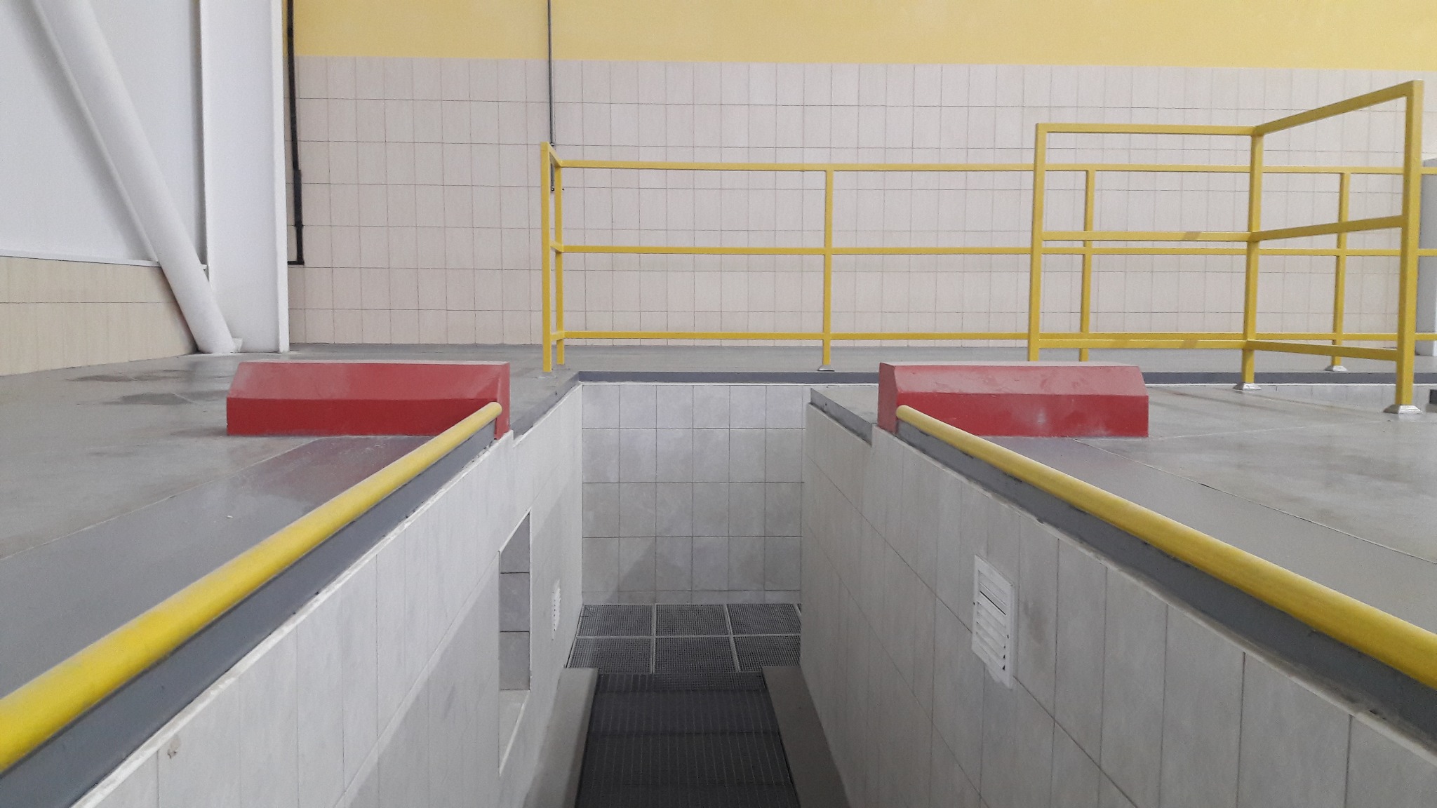 Wnętrze hali przemysłowej z zabezpieczonym kanałem rewizyjnym, wykończonym płytkami i kratą pomostową, z żółtymi barierkami i czerwonymi odbojnikami.