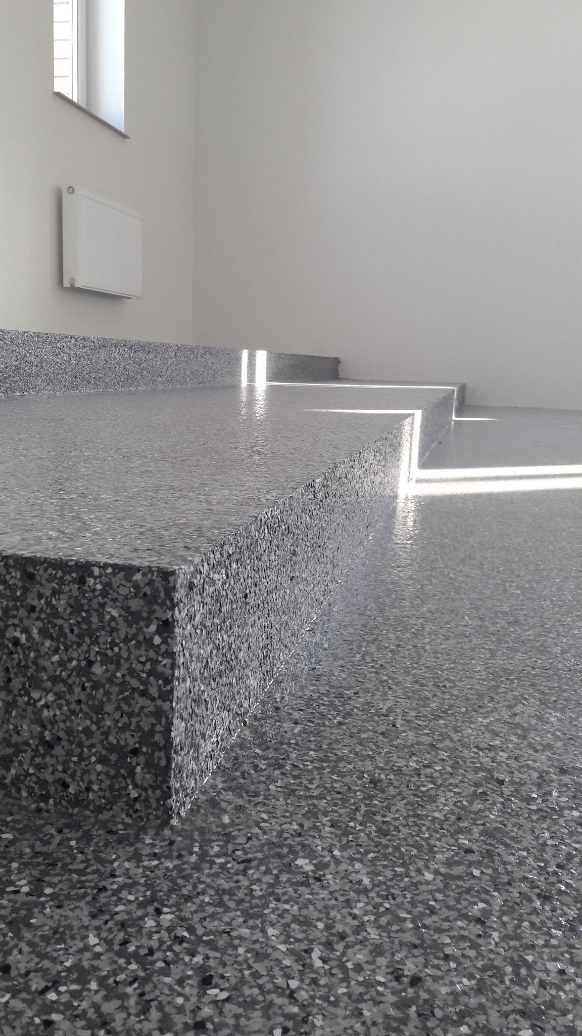 Szara, błyszcząca podłoga z żywicy z wyraźną fakturą, oświetlona promieniami słońca wpadającymi przez okno, widoczne schodkowe podwyższenia podłogi.