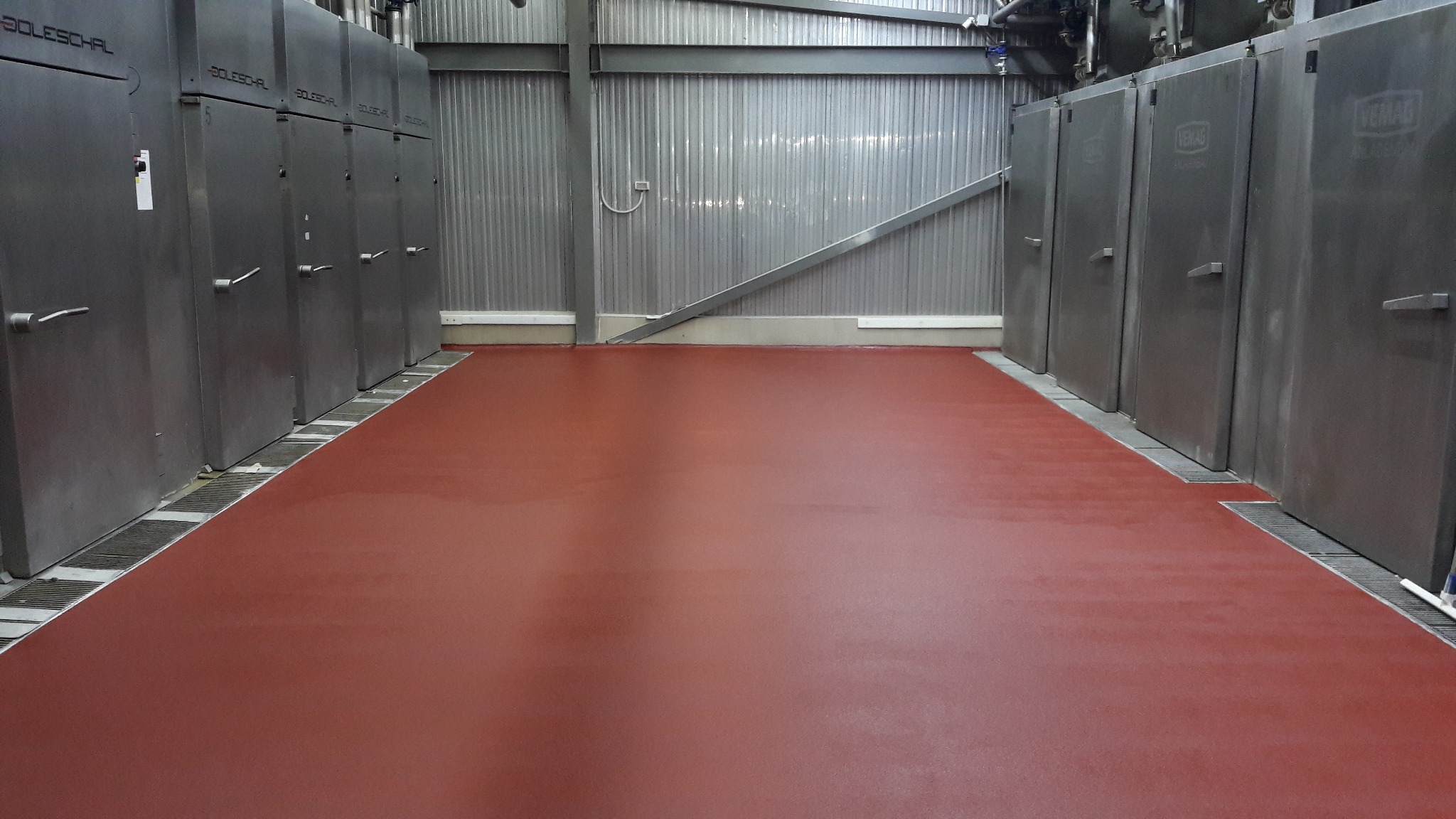 Czerwona, gładka podłoga w hali przemysłowej z metalowymi szafami po bokach i kratkami odpływowymi wzdłuż ścian.