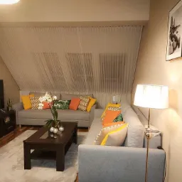 Jasne, delikatne zasłony w skosie okna mansardowego, dopełniające aranżację salonu z szarymi sofami i kolorowymi poduszkami.