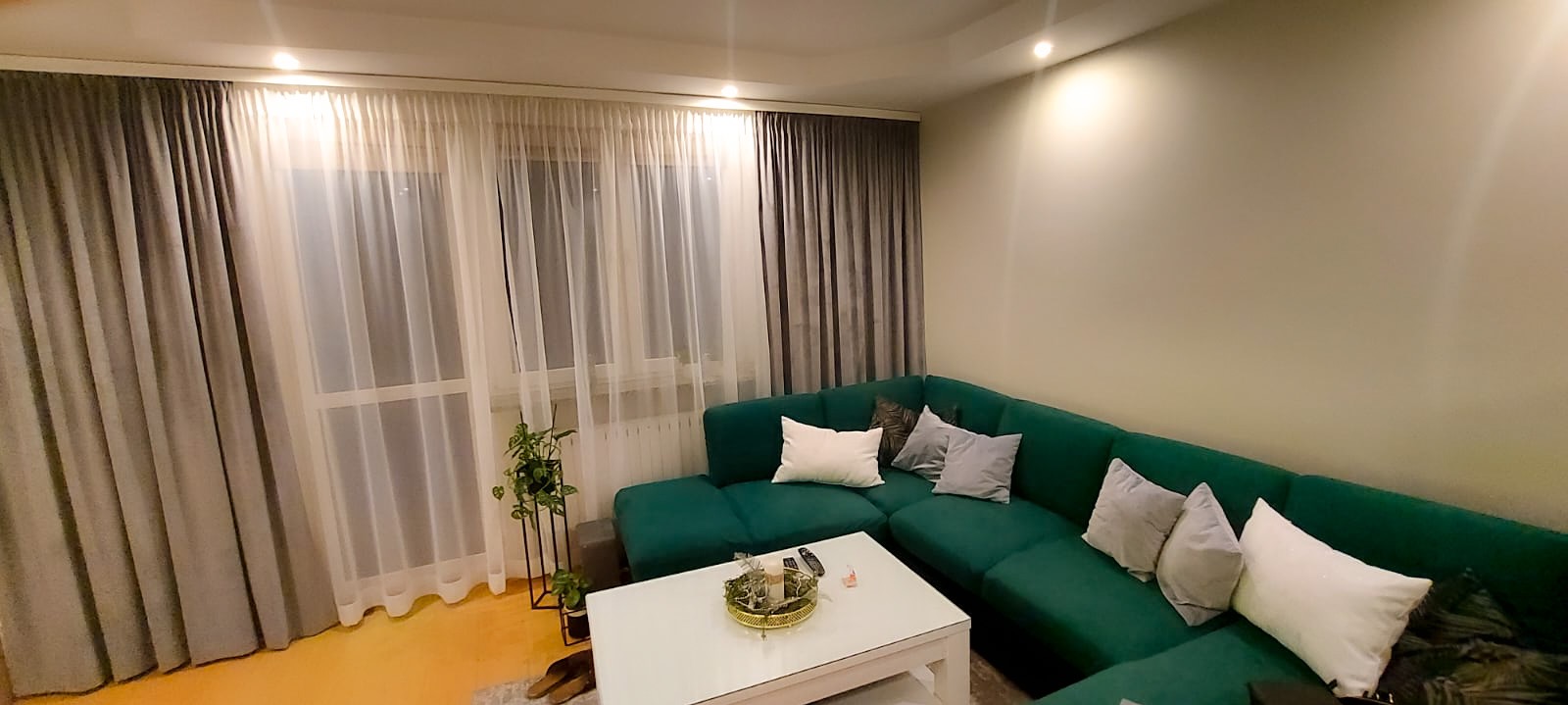 Szare i białe zasłony w nowoczesnym salonie z zieloną sofą narożną i białym stolikiem kawowym.