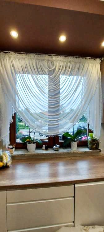 Delikatna, biała firanka z koralikami zdobiąca okno, na parapecie doniczki z roślinami, widok na zewnątrz przez okno.