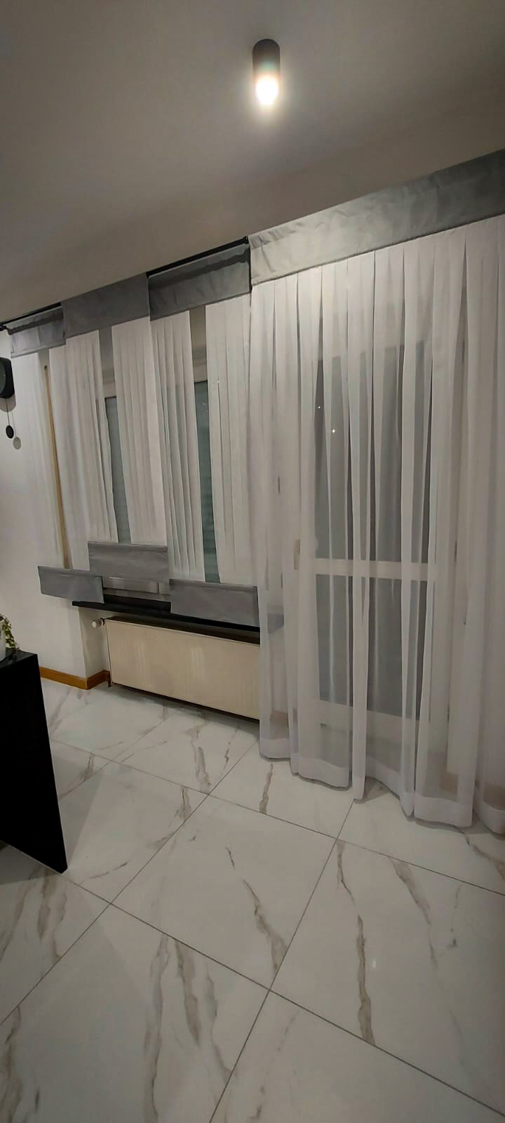 Delikatne, białe, półprzezroczyste zasłony z szarymi karniszami, wiszące w nowoczesnym wnętrzu z marmurową podłogą i grzejnikiem pod oknem.