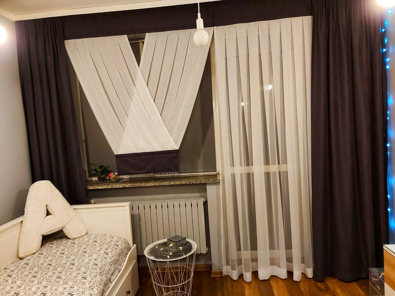 Pokój dziecięcy z oknem ozdobionym brązowymi zasłonami i białymi, zwiewnymi firanami. Na parapecie doniczka z rośliną. Obok okna stoi białe łóżko z poduszką w kształcie litery A i stolik nocny...