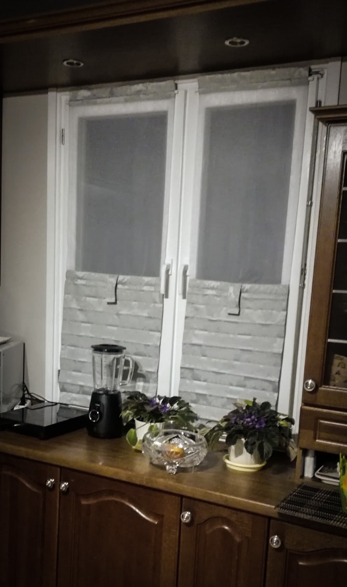 Dwu-częściowe szare zasłony panelowe zawieszone na białym oknie, widoczne z blatem kuchennym, na którym stoją blender, kwiaty w doniczkach i kryształowa misa.