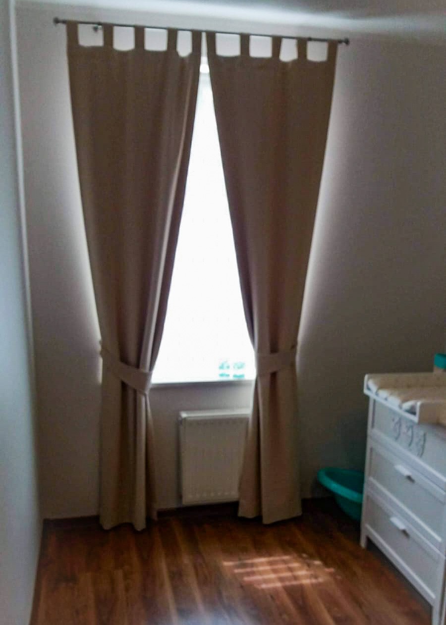 Beżowe zasłony z podwiązkami w pokoju dziecięcym, widok na okno i białą komodę z przewijakiem.