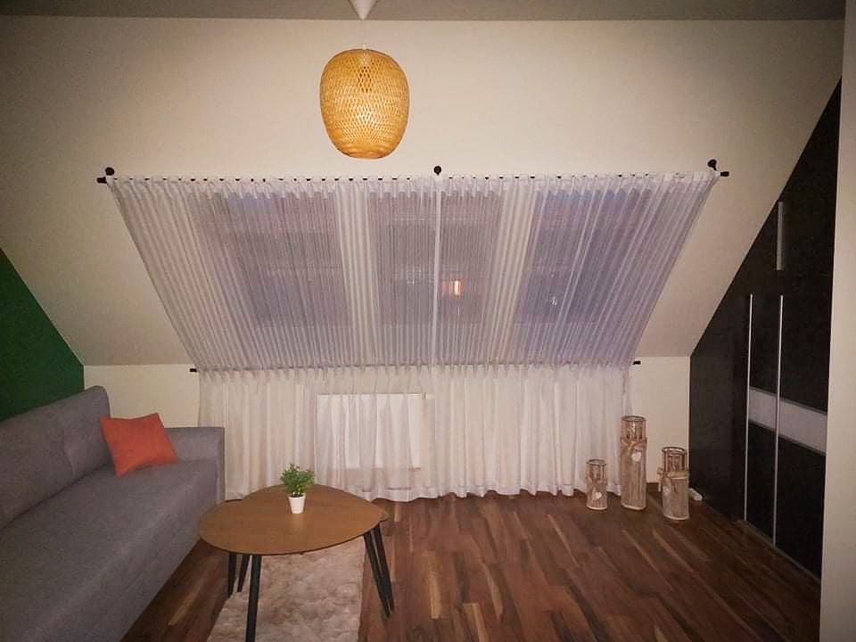 Jasne, transparentne zasłony w oknie na poddaszu, dopasowane do skośnego sufitu, z widocznym szarym narożnikiem, drewnianym stolikiem kawowym i dekoracyjnymi wazonami na podłodze.