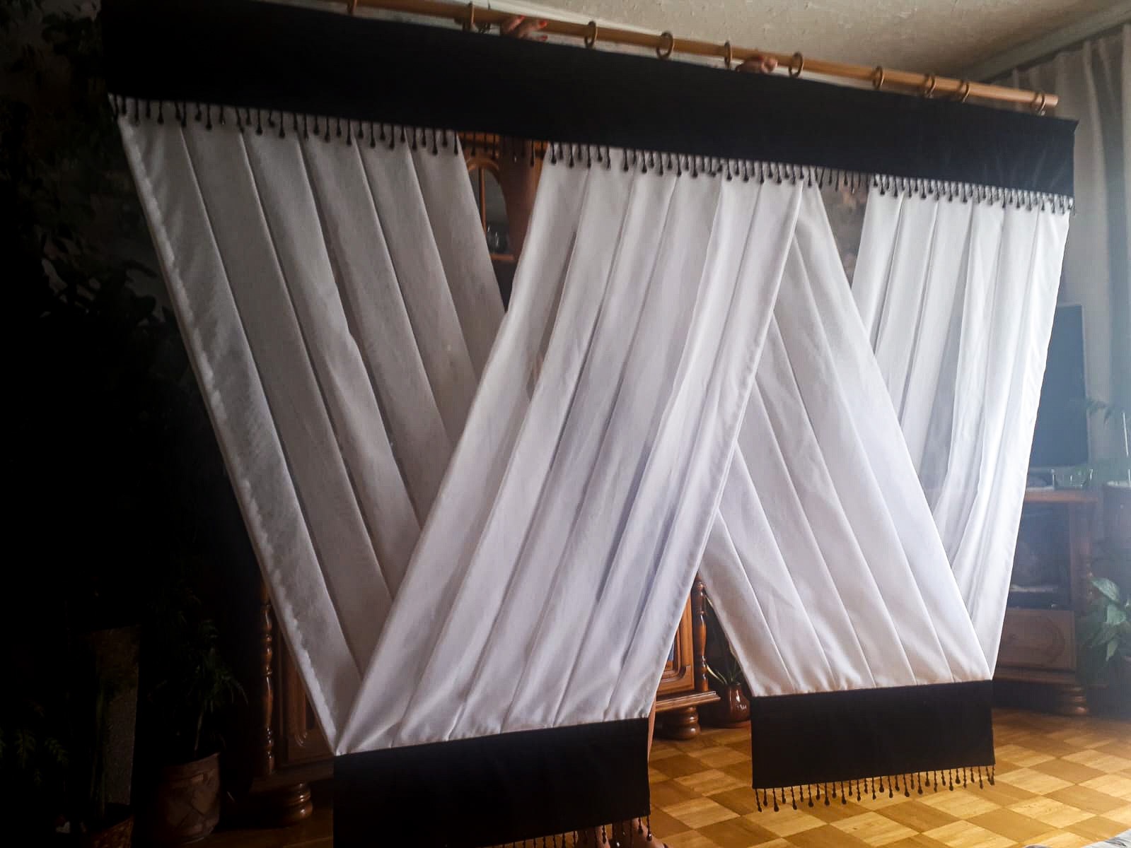 Draperie okienne z białego materiału z czarnym wykończeniem i dekoracyjnymi frędzlami, zawieszone na drewnianym karniszu w klasycznym wnętrzu.