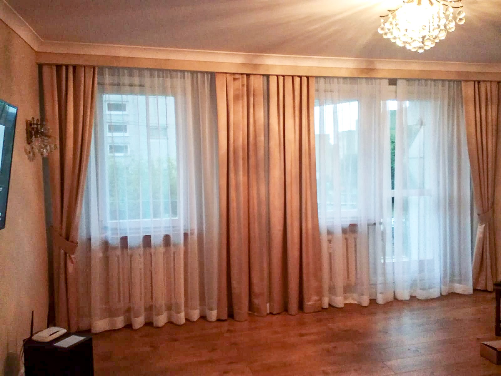 Salon z trzema oknami udekorowanymi długimi, beżowymi zasłonami i białymi firanami, oświetlony kryształowym żyrandolem.