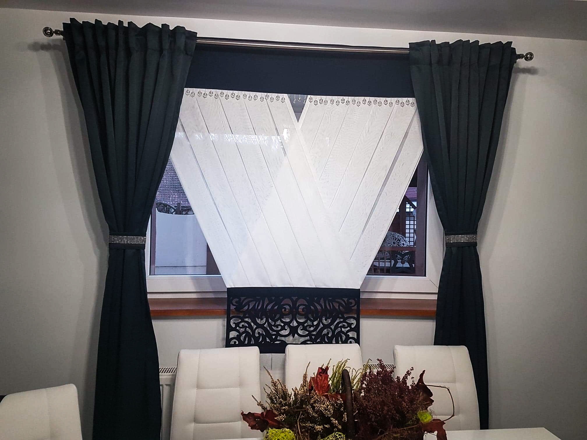 Eleganckie, ciemnozielone zasłony z ozdobnym pasem cyrkonii, połączone z białą, dekoracyjną firaną w geometryczne wzory, tworzące spójną aranżację okienną nad stołem z bukietem jesiennych kwiatów.
