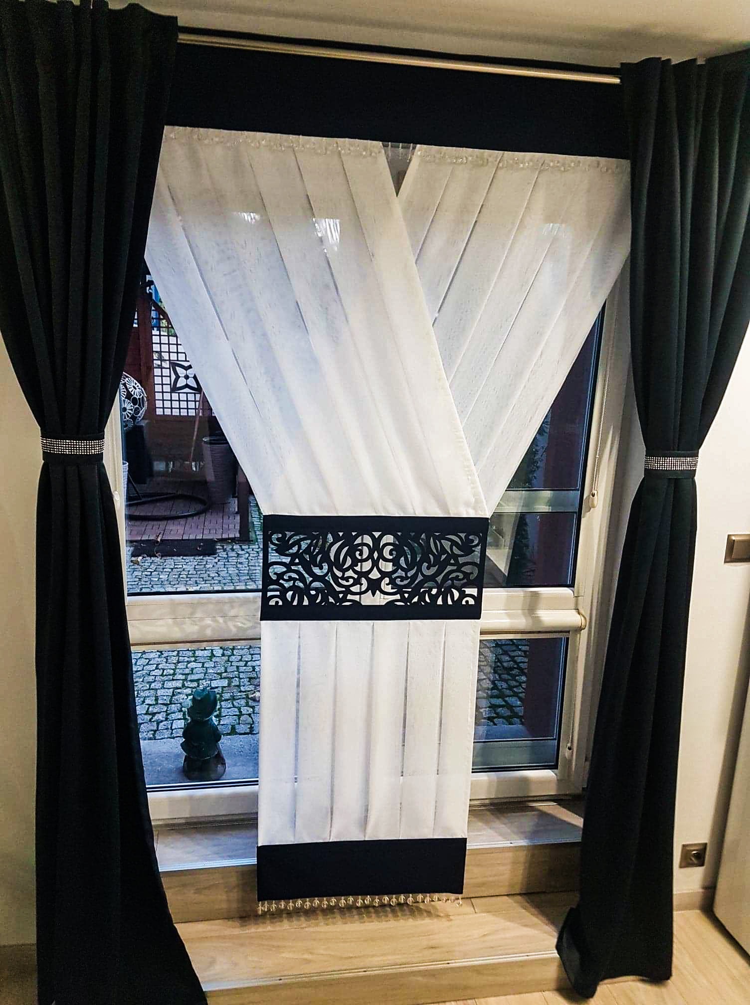 Biała, transparentna zasłona z czarnym, ażurowym pasem i czarnym dołem z frędzlami, otoczona ciężkimi, czarnymi zasłonami z ozdobnymi upięciami na tle okna z widokiem na ogród z kamienną ścieżką...