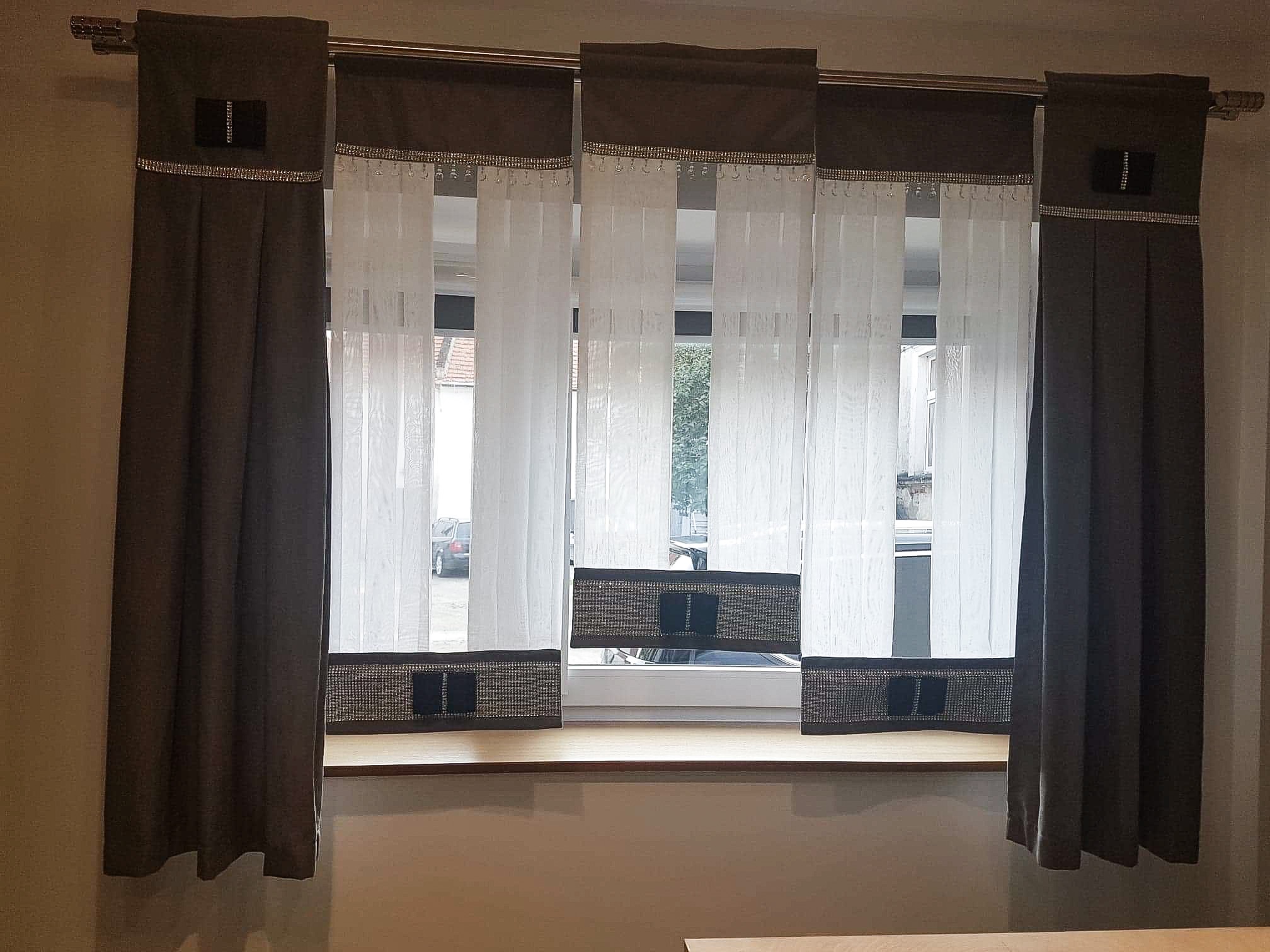 Kompozycja okienna z brązowych zasłon bocznych z dekoracyjnym paskiem i etykietą oraz białych, pionowych żaluzji z brązowymi akcentami u góry i na dole, wykończonych błyszczącą taśmą.