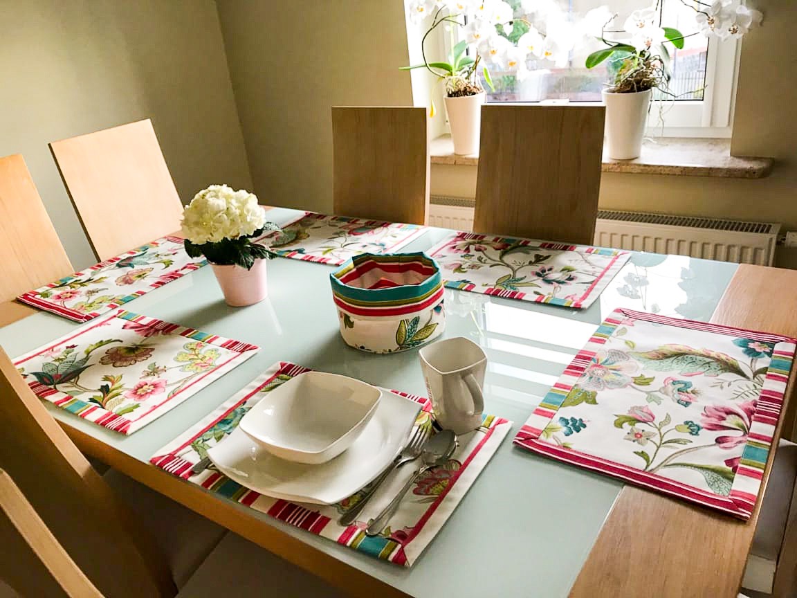 Elegancko nakryty stół z białym serwisem, kwiatami w różowej doniczce i pasującymi podkładkami oraz koszyczkiem w kwiatowy wzór z czerwonymi paskami.