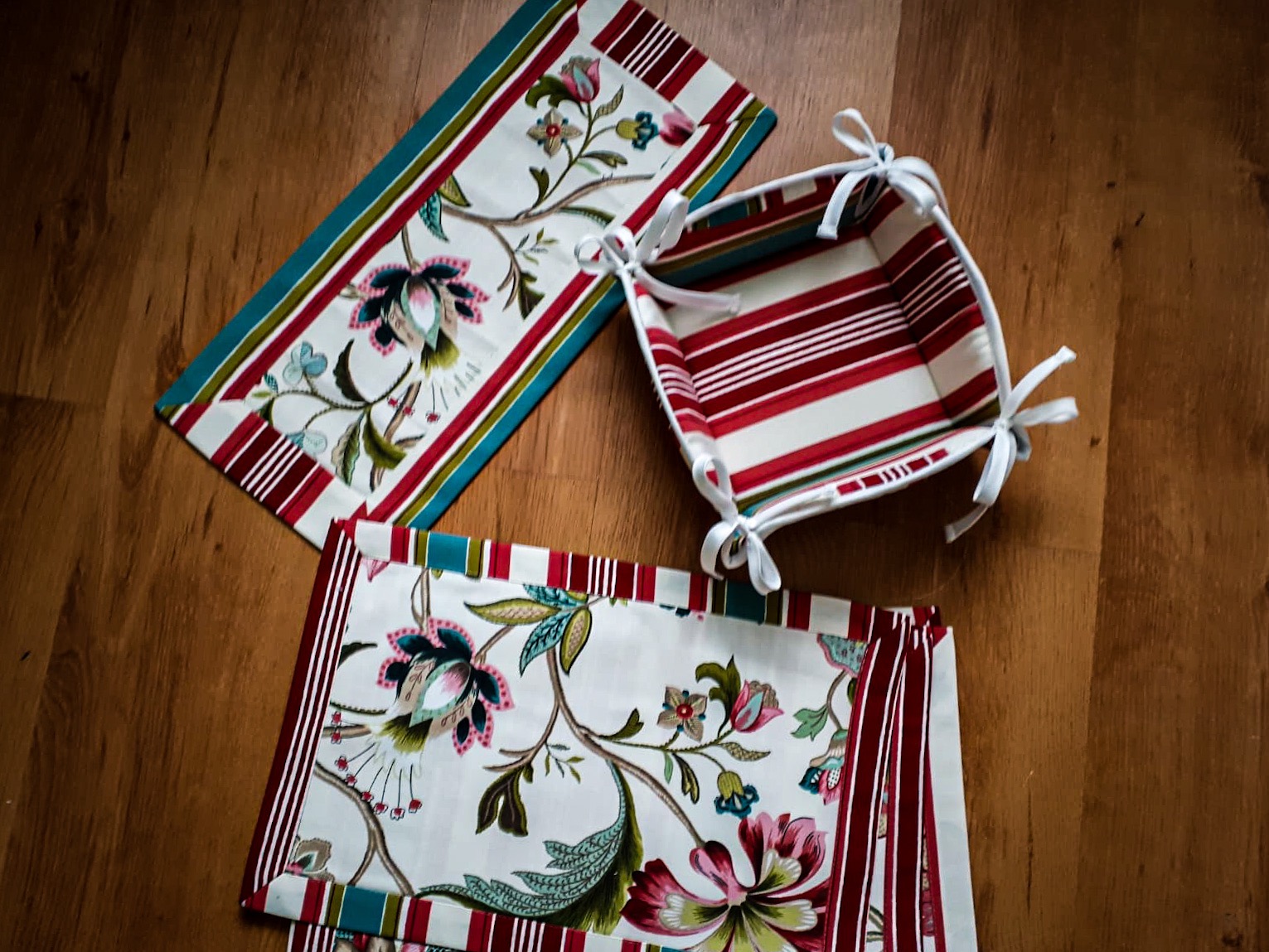 Komplet tekstylny: bieżnik z motywem kwiatowym, podkładki i koszyk z czerwono-białymi paskami na drewnianym tle.