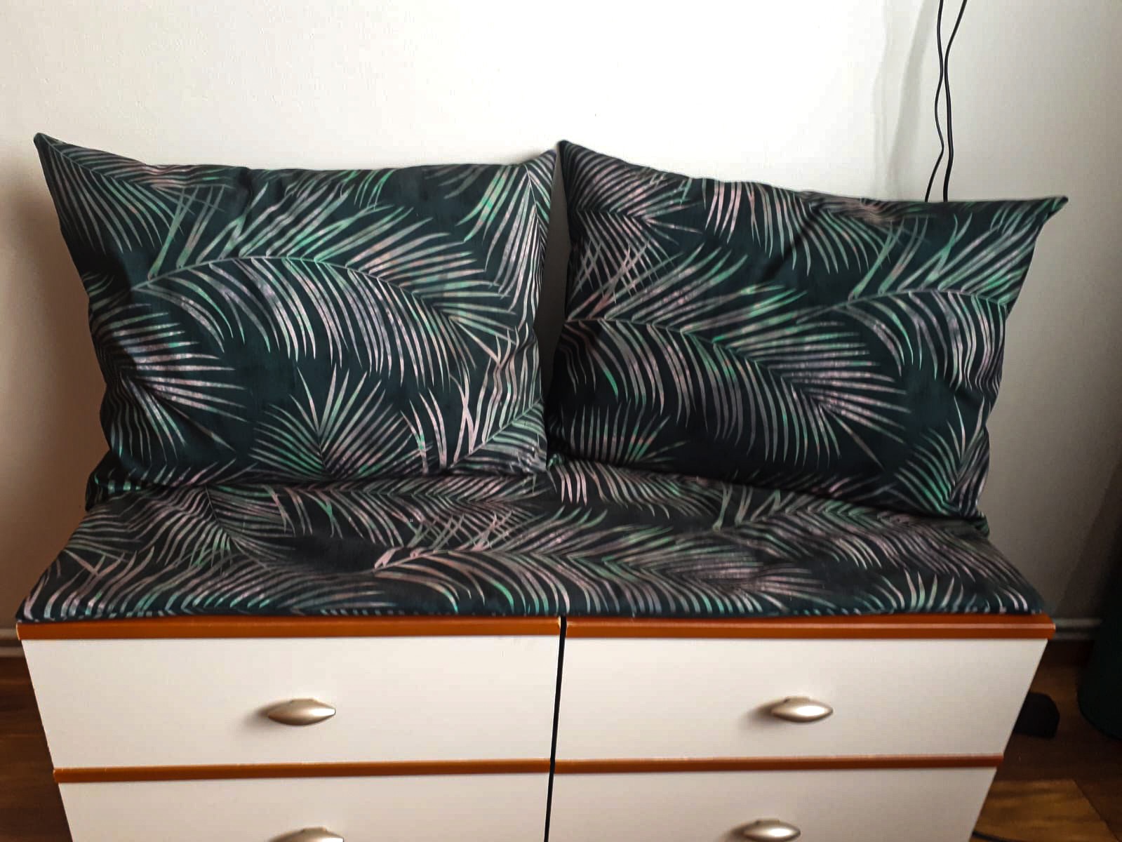 Dwa kwadratowe, ciemnozielone poduszki z motywem liści palmowych, leżące na pokrowcu z tego samego materiału, umieszczonym na białej komodzie z czterema szufladami.