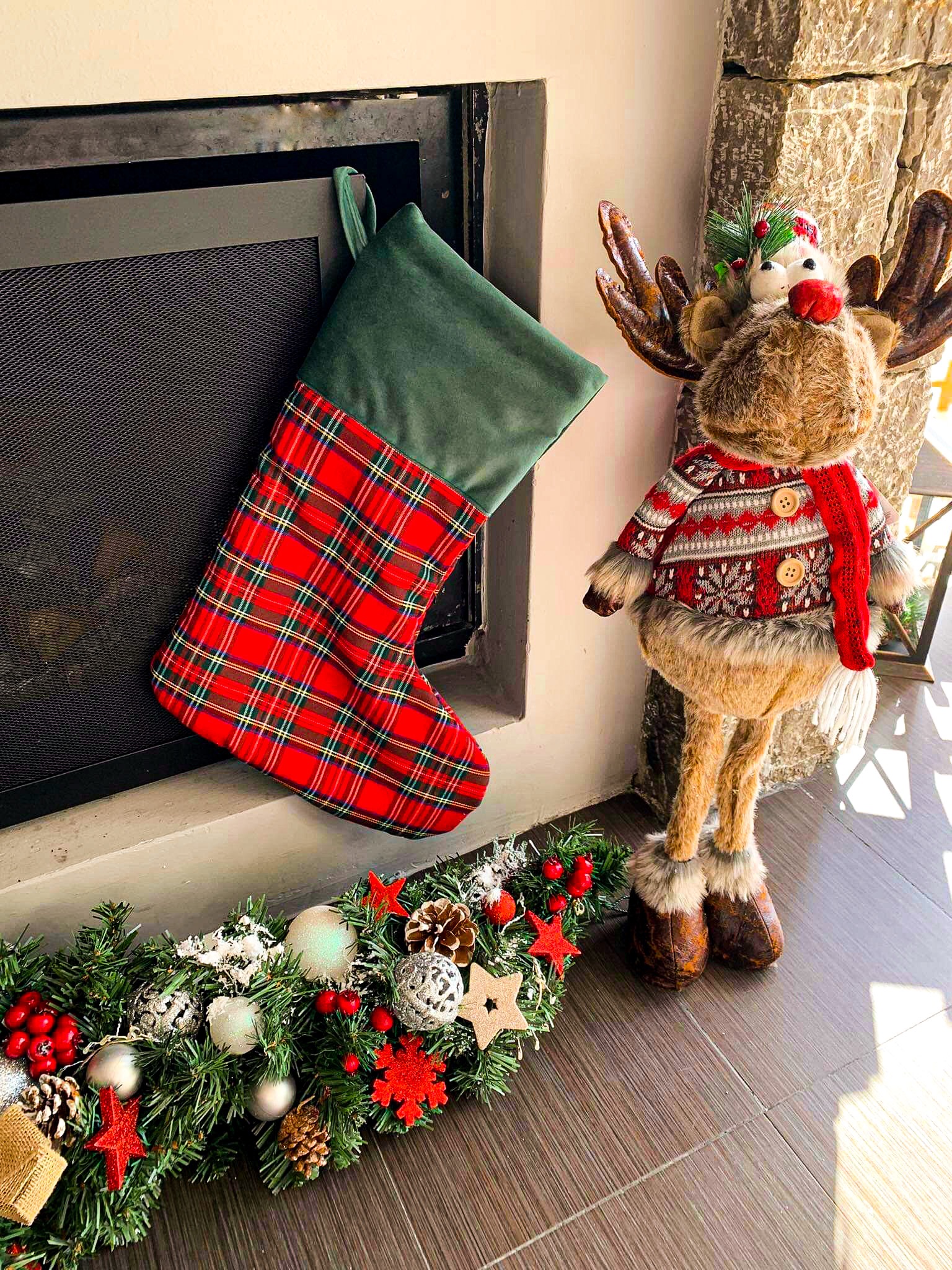 Świąteczna dekoracja kominka z zielono-czerwonym, kraciastym workiem na prezenty, pluszowym reniferem w swetrze i girlandą z bombkami.