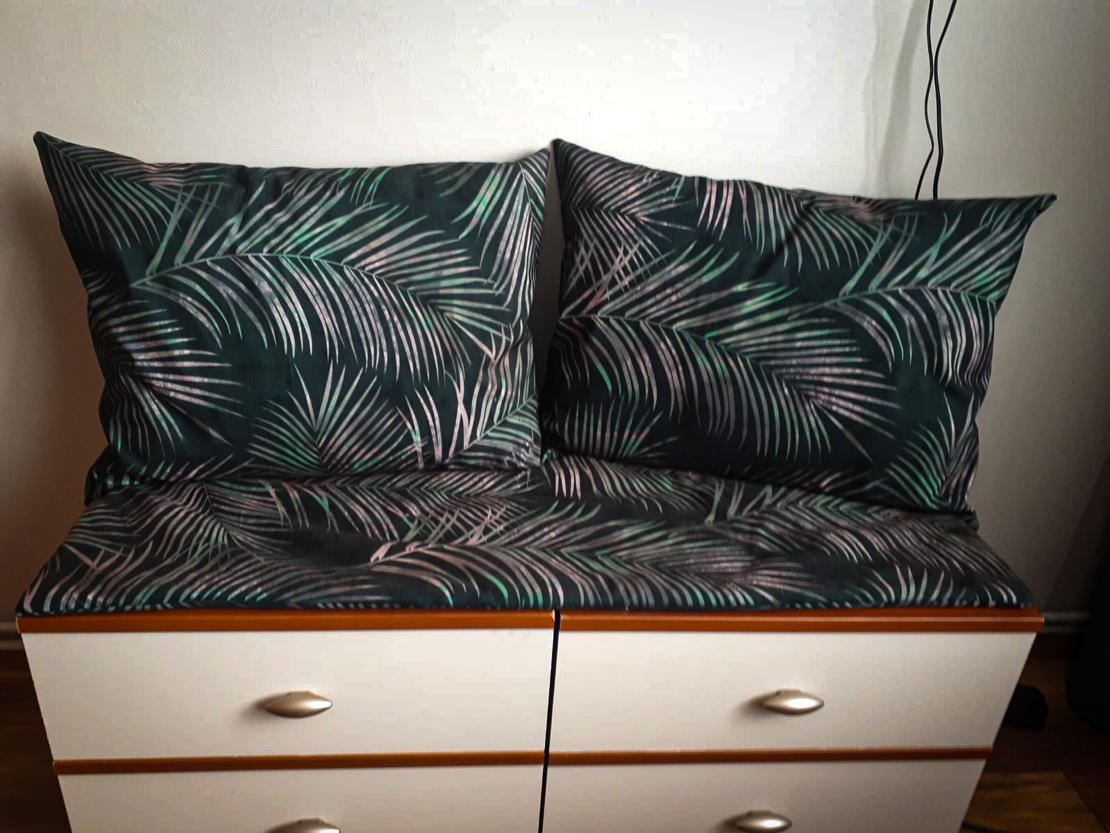 Dwie poduszki i siedzisko z ciemnej tkaniny w motyw liści palmowych na białej komodzie z czterema szufladami.