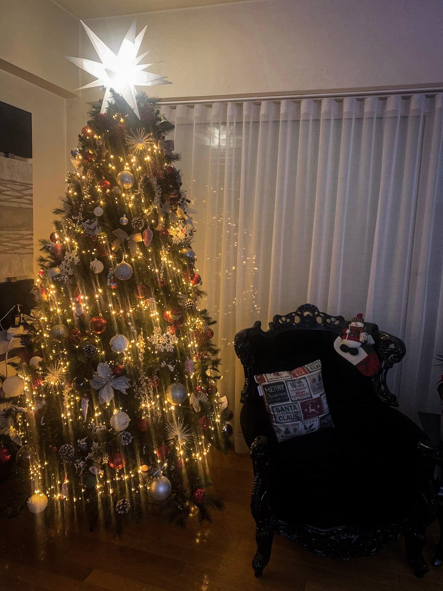 Oświetlona choinka z białymi i czerwonymi ozdobami, zwieńczona świecącą gwiazdą, obok czarne, zdobione krzesło z poduszką świąteczną i skarpetą św. Mikołaja, na tle białych, lekko prześwitujących...