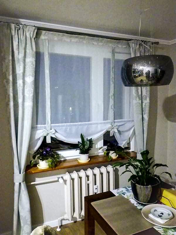 Jasne, wzorzyste zasłony okienne z dekoracyjnymi kokardami, w tle okno z roślinami doniczkowymi na parapecie, widoczny element grzejnika i lampa sufitowa.