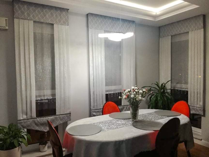 Eleganckie, pionowe żaluzje z szarą, wzorzystą falbaną w jadalni z okrągłym stołem i pomarańczowymi krzesłami.