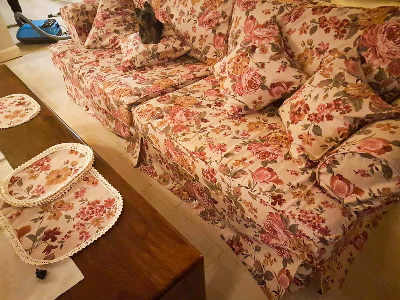 Komplet pokrowców w kwiaty na sofę i poduszki, z dopasowanymi serwetami na stoliku kawowym, na sofie siedzi mały pies.