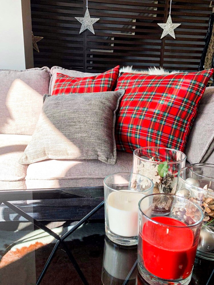 Dekoracyjny salon z sofą z poduszkami w kratę i szarą, szklany stolik kawowy z ozdobnymi świecami w szklanych naczyniach i gwiazdami zawieszonymi na górze.