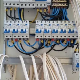 Rozdzielnia elektryczna z widocznymi bezpiecznikami, przewodami oznaczonymi etykietami L1, L2, L3, oraz listwami zaciskowymi, zamontowana na ścianie z widocznymi ubytkami tynku.