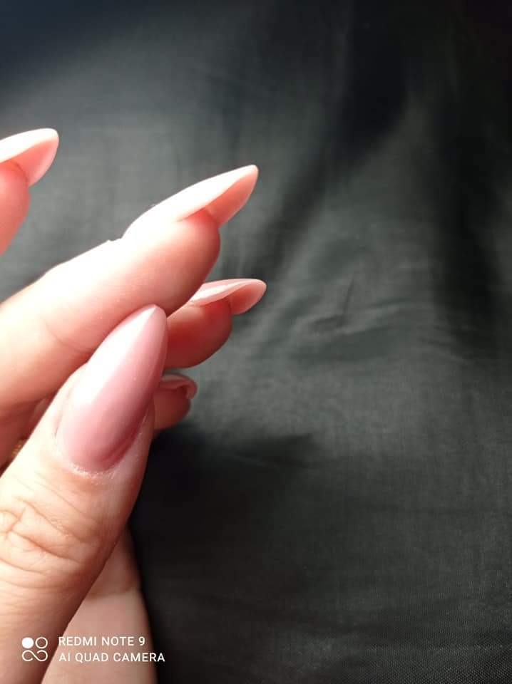 Dłoń z przedłużonymi, migdałowymi paznokciami w odcieniu pudrowego różu na ciemnym tle.