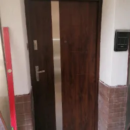 Ciemnobrązowe drzwi wejściowe z pionowym, srebrnym panelem, zamontowane w nierównym otworze, poziomica w tle, widoczny fragment ceglanej ściany.