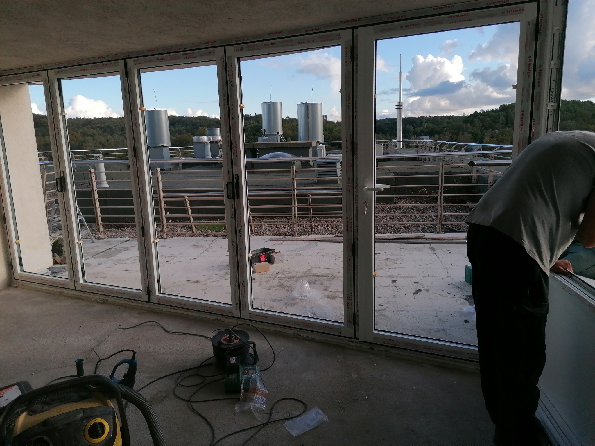 Montaż dużych, przesuwnych okien balkonowych PCV z widokiem na zewnątrz, narzędzia pracy na podłodze, w tle zieleń i elementy infrastruktury miejskiej.