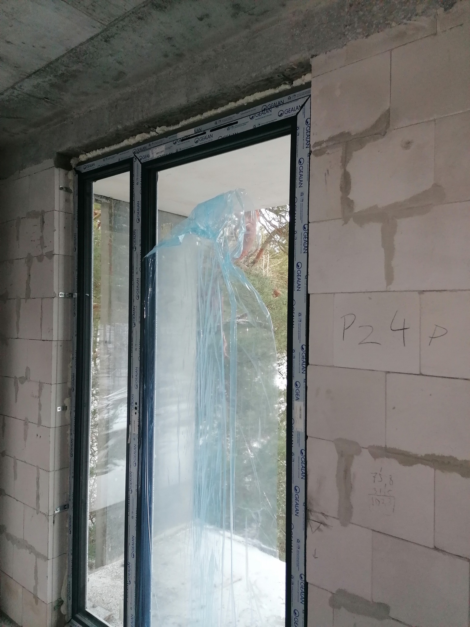 Montaż okna PCV w surowym stanie, zabezpieczone folią, widoczne pianowanie i bloczki betonowe z notatkami.