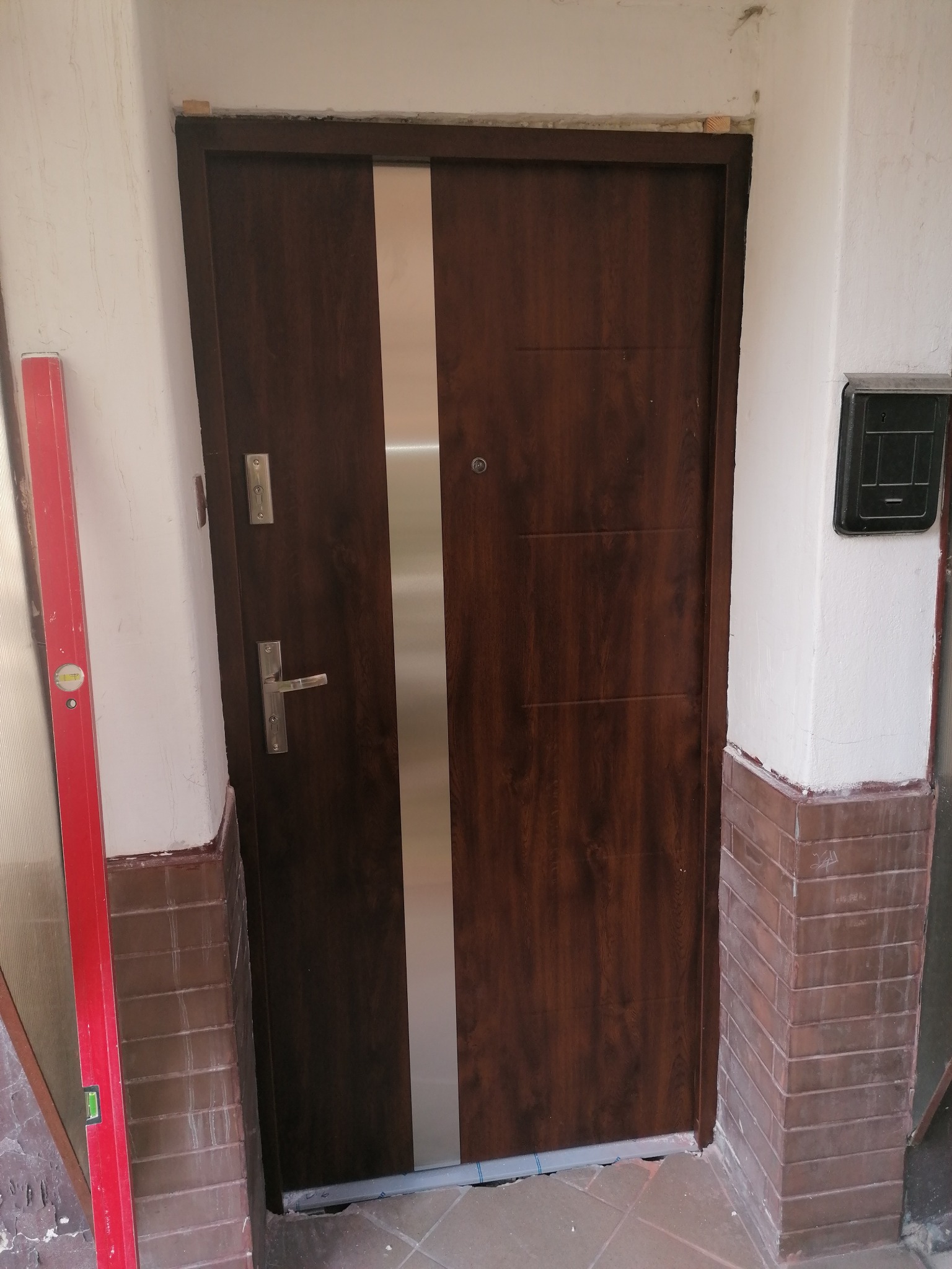 Ciemnobrązowe drzwi wejściowe z pionowym, srebrnym panelem, zamontowane w nierównym otworze, poziomica w tle, widoczny fragment ceglanej ściany.