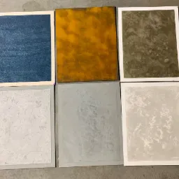 Sześć próbek kolorów i faktur farb lub tynków dekoracyjnych na kartonach, odcienie niebieskiego, żółtego, szarego i białego, ułożone na szarym podłożu.