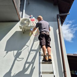 Elektryk Zielonka - Serwisant na drabinie podłącza zestaw manometrów do jednostki zewnętrznej klimatyzatora VIVAX, zamontowanej na jasnej elewacji budynku, w słoneczny dzień.