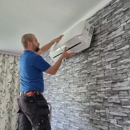 Elektryk Zielonka - Technik w niebieskiej koszulce i roboczych spodniach z narzędziami na pasku instaluje biały klimatyzator ścienny na tle szarej tapety imitującej kamień, obok okna z zielonymi zasłonami.