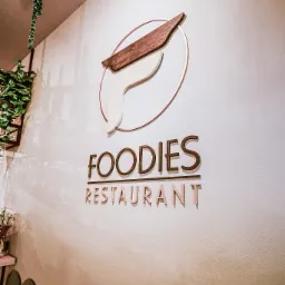 Logo restauracji Foodies na białej ścianie z dekoracjami roślinnymi i rzędem zielonych krzeseł w tle.