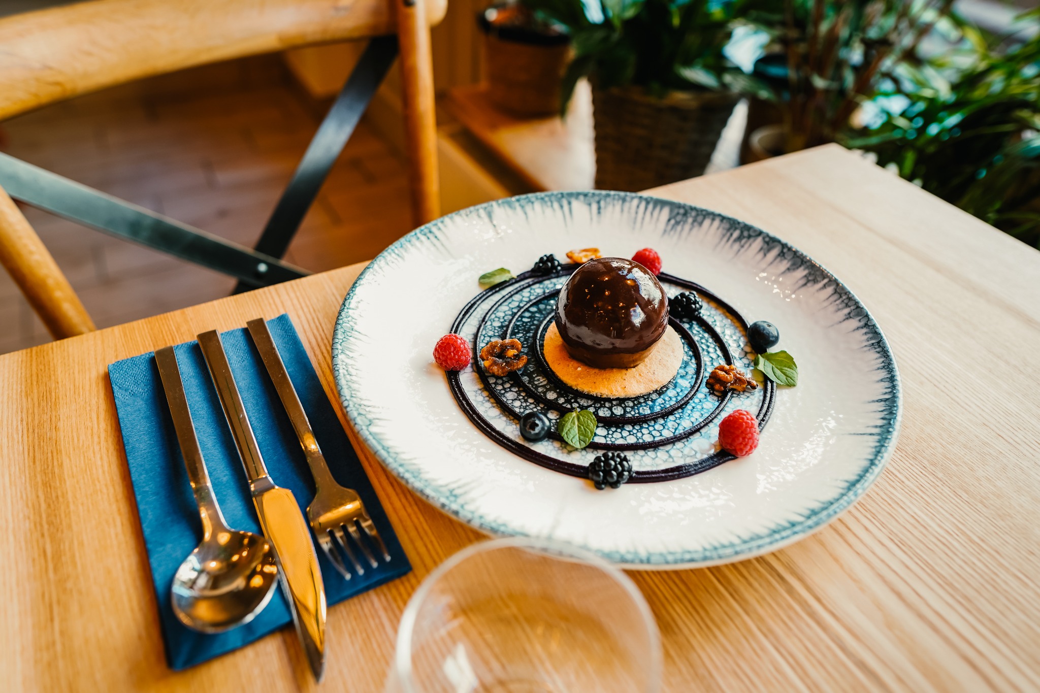 Elegancko podany deser: czekoladowa kula na ciastku, otoczona sosem, malinami, jeżynami, borówkami i orzechami na talerzu z niebieskim wzorem, obok niebieska serwetka ze złotymi sztućcami,...