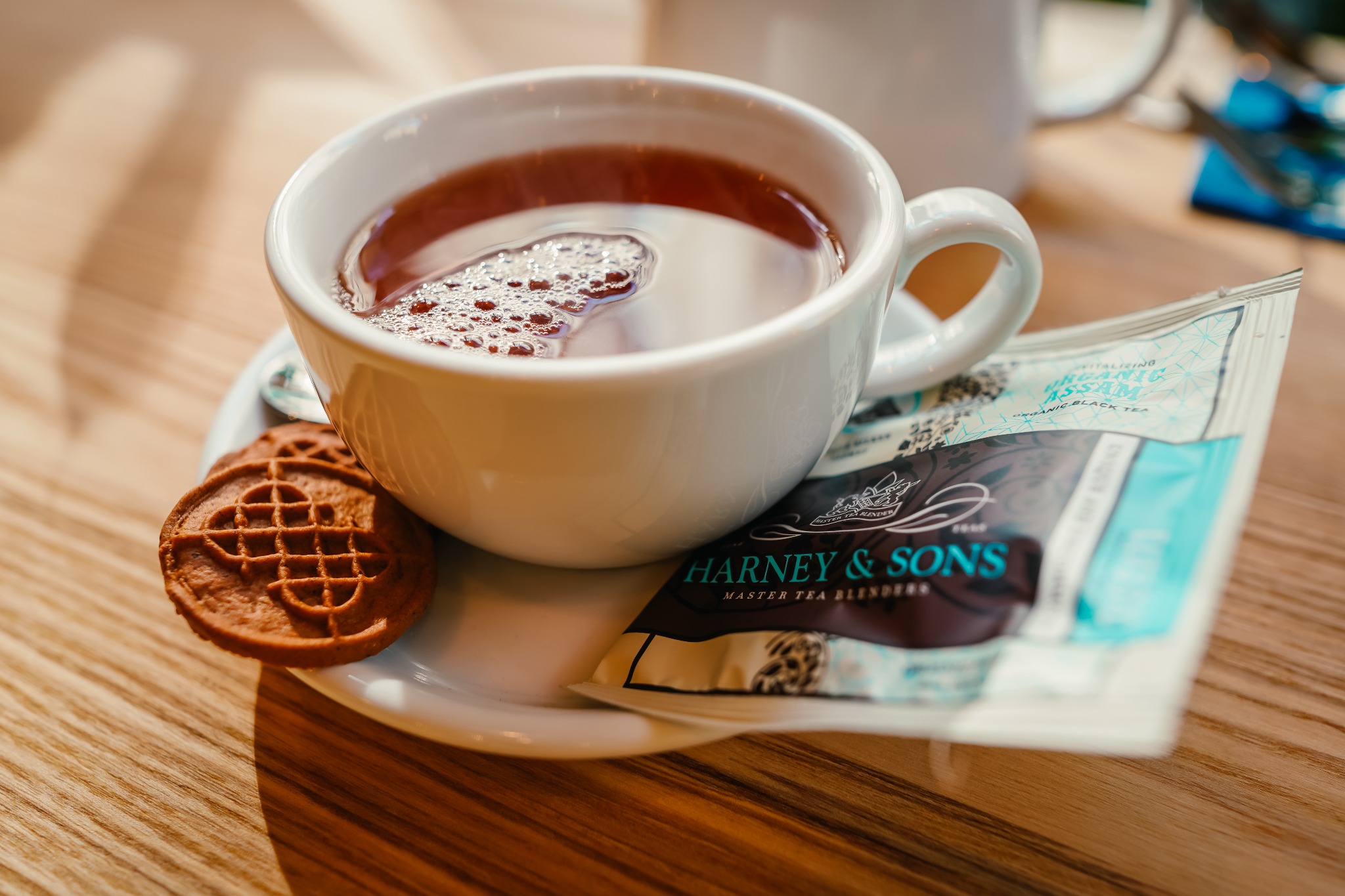 Biała filiżanka z herbatą Assam, z widocznymi bąbelkami na powierzchni, stoi na spodku obok dwóch ciasteczek z wzorem kratki i saszetki herbaty Harney & Sons na drewnianym stole.