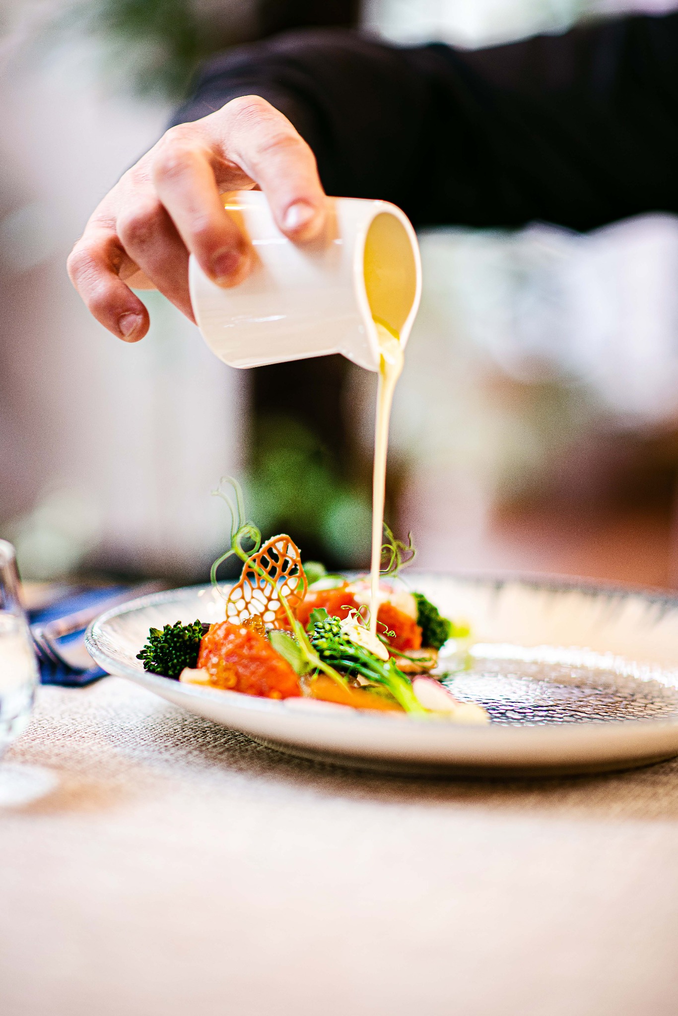 Szef kuchni polewa sosem danie z brokułami, pomidorami i dekoracją z jadalnej koronki na talerzu o fakturze szkła