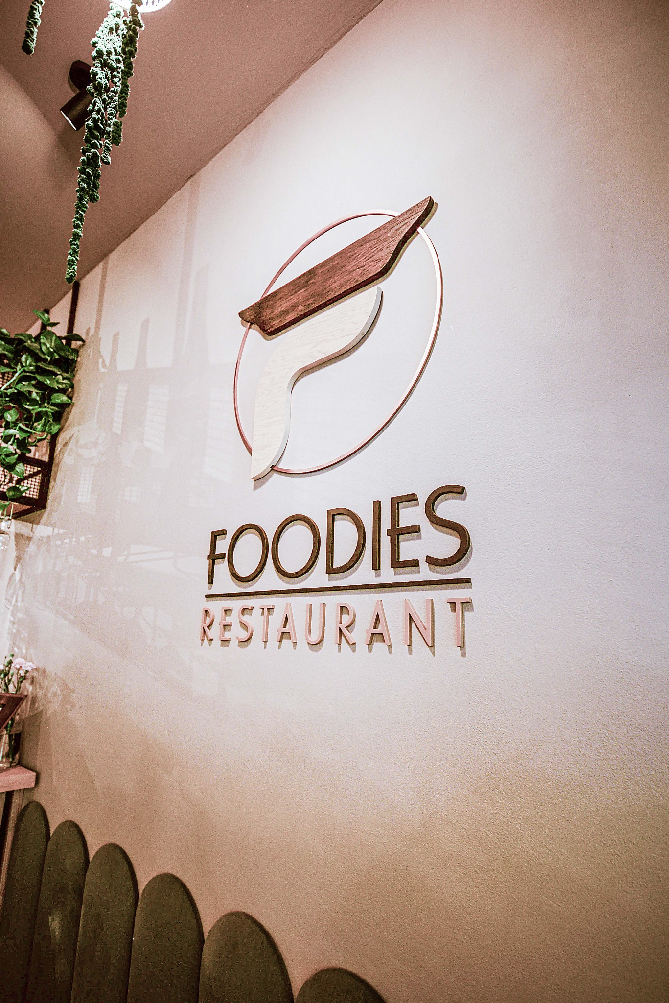 Logo restauracji Foodies na białej ścianie z dekoracjami roślinnymi i rzędem zielonych krzeseł w tle.