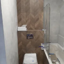 Łazienka w trakcie remontu: brązowe płytki ułożone w jodełkę na ścianie za toaletą i nad nią, białe płytki po lewej stronie, wanna z zamontowanym parawanem w trakcie zabezpieczania folią malarską...