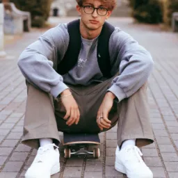 Młody mężczyzna w okularach siedzi na deskorolce na brukowanej ścieżce, ubrany w szarą bluzę z napisem, khaki spodnie i białe sneakersy, z czarnym plecakiem na plecach, patrząc wprost w obiektyw.