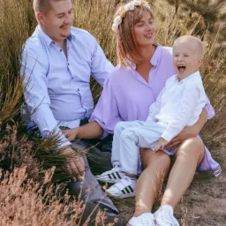 Uśmiechnięta rodzina siedzi w trawie: tata w błękitnej koszuli, mama z wiankiem na głowie w liliowej sukience trzyma na kolanach roześmianego synka w białej koszuli i jasnych spodniach.