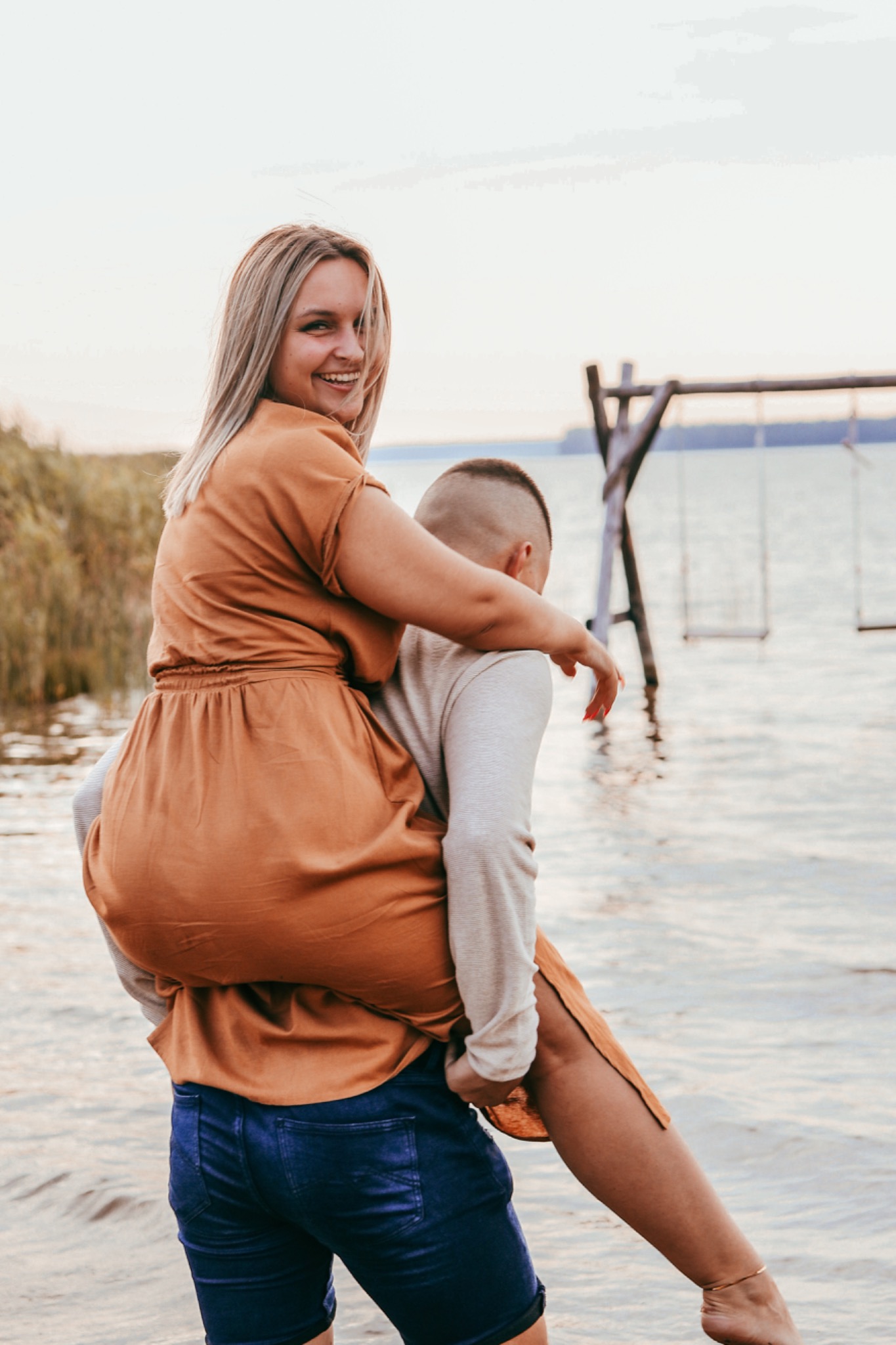Uśmiechnięta blondynka w pomarańczowej sukience na barana u mężczyzny na brzegu jeziora, w tle huśtawki i zachodzące słońce.